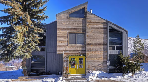 A modern wooden house with a unique design, featuring a bright yellow door and surrounded by snow and trees.